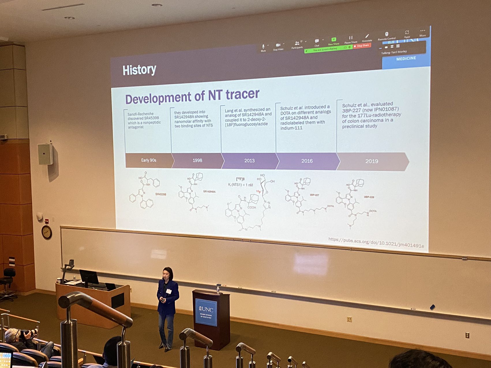 woman standing at the front of an auditorium with "Development of NT Tracer" written on a large PowerPoint screen